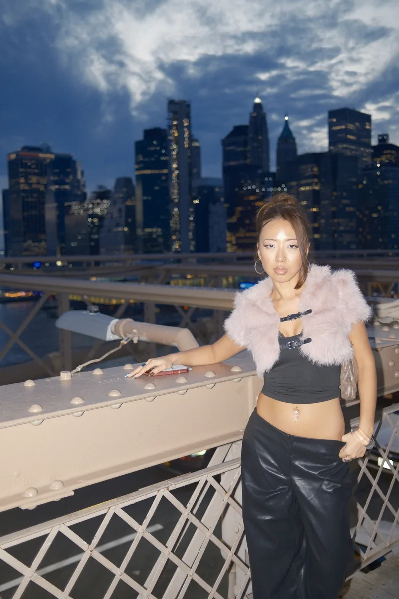 A woman in a pink fur shrug and black leather trousers leans on the Brooklyn Bridge railing and looks straight at the camera, the Lower Manhattan skyline lit up behind her at blue hour