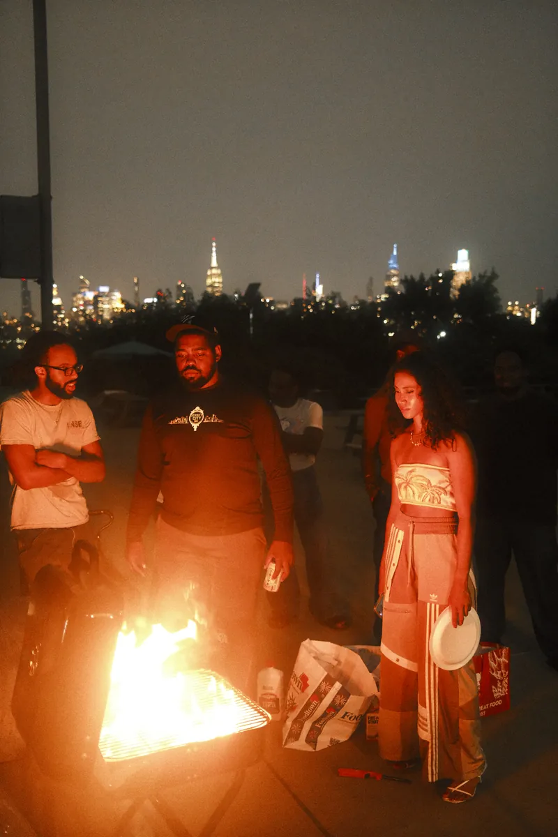 Friends around a flaming grill at night with the Manhattan skyline behind