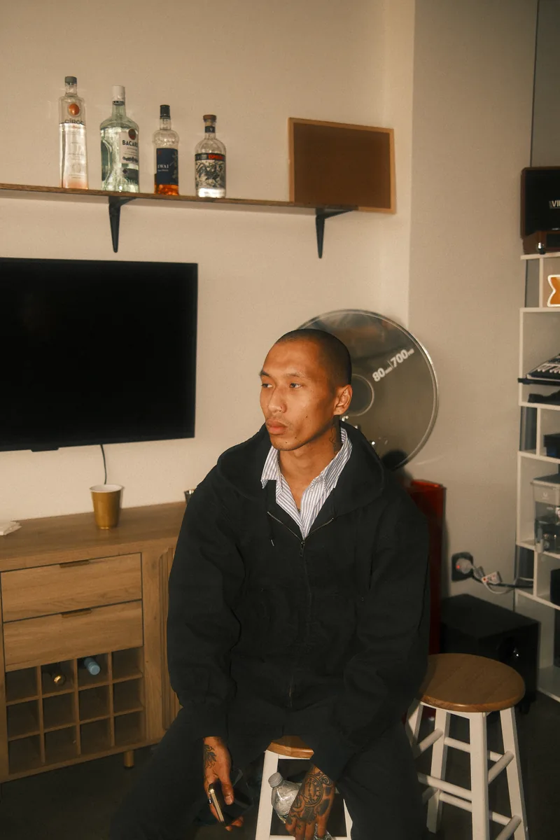 A friend with a shaved head and tattooed hands sits on a stool at an OTL recording session, a giant CD clock and a shelf of bottles behind him