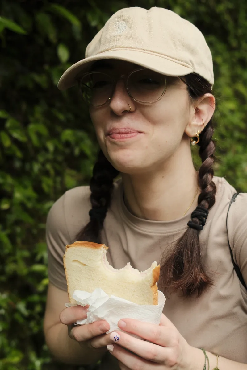 A friend in a cap and round glasses mid-bite of a sandwich against deep green leaves