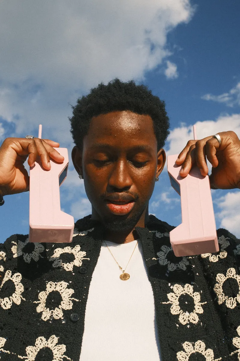 Portrait in a crochet shirt holding two pink brick phones against a blue sky
