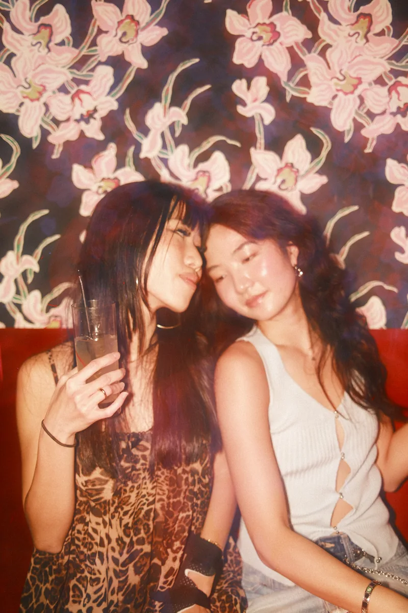Two friends leaning into each other on a red banquette under pink floral wallpaper